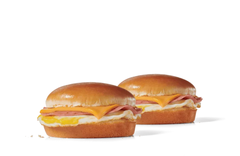 Two breakfast sandwiches on a transparent background, each featuring a golden-brown bun, slices of ham, melted cheese, and a fluffy egg layer.