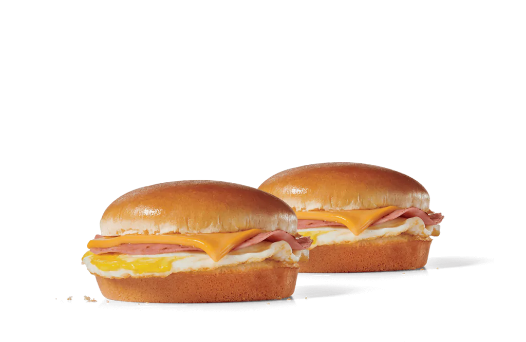 Two breakfast sandwiches on a transparent background, each featuring a golden-brown bun, slices of ham, melted cheese, and a fluffy egg layer.