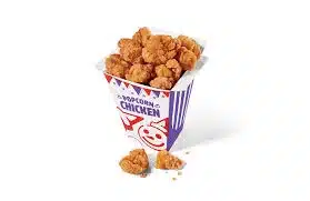 A festive carton filled to the brim with crispy golden popcorn chicken. The pieces are bite-sized and uniformly cooked, presented in a purple and white striped carton adorned with playful graphics, a few pieces scattered around the base on a light background.