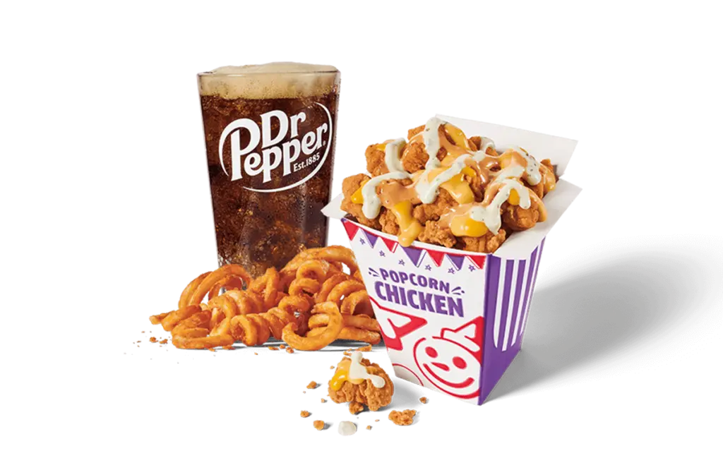 A box of sauced and loaded popcorn chicken topped with white creamy sauce, paired with a portion of curly fries, alongside a glass of Dr Pepper, set against transparent background.