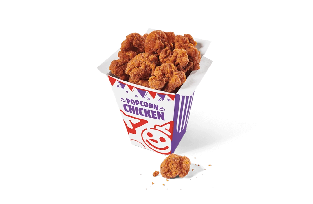 A carton filled with spicy popcorn chicken pieces, presented on transparent background. The chicken is deep-fried to a crispy golden brown, and the colorful carton is adorned with playful graphics and purple stripes, with some crumbs scattered beside it.