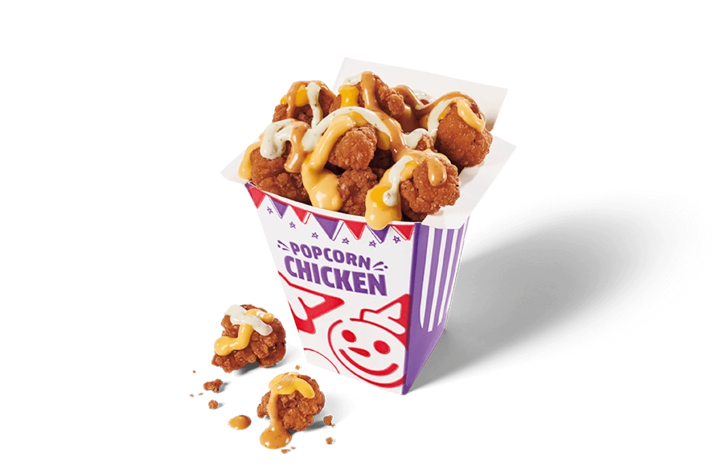 A vibrant carton overflowing with spicy popcorn chicken, generously topped with swirls of yellow and white sauces. The nuggets are golden-brown and crispy, some spilling out onto the transpparent background, with crumbs visible around the carton.