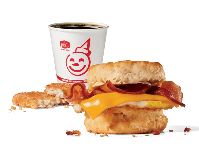 A delicious breakfast combo featuring a freshly baked biscuit sandwich filled with crispy bacon, melted cheese, and a fluffy egg, accompanied by a hash brown patty and a hot coffee in a branded Jack in the Box cup, all set against a transparent background.