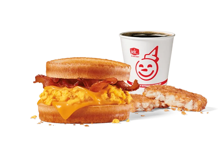 An appetizing image featuring Jack in the Box's Bacon, Egg & Cheese Scrambler Combo. This breakfast sandwich consists of scrambled eggs, melted cheese, and crispy bacon stacked between two golden muffin halves. Accompanying the sandwich is a hash brown and a coffee cup with the smiling Jack in the Box logo, all presented on a transparent backdrop to enhance the visual appeal of the meal.