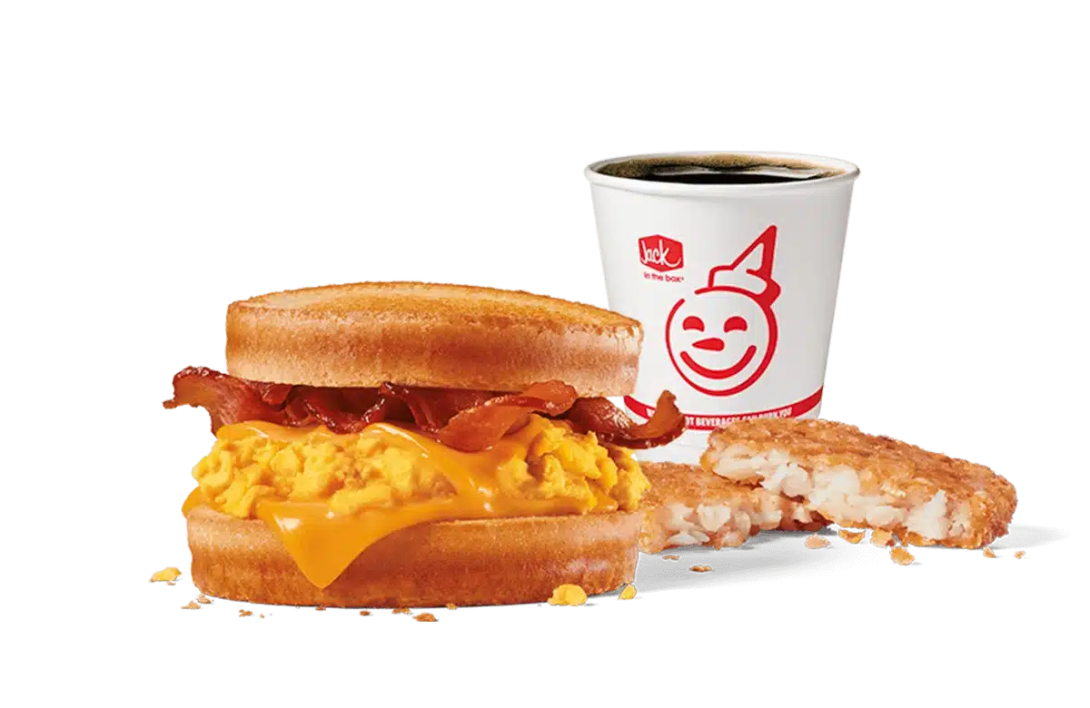 An appetizing image featuring Jack in the Box's Bacon, Egg & Cheese Scrambler Combo. This breakfast sandwich consists of scrambled eggs, melted cheese, and crispy bacon stacked between two golden muffin halves. Accompanying the sandwich is a hash brown and a coffee cup with the smiling Jack in the Box logo, all presented on a transparent backdrop to enhance the visual appeal of the meal.