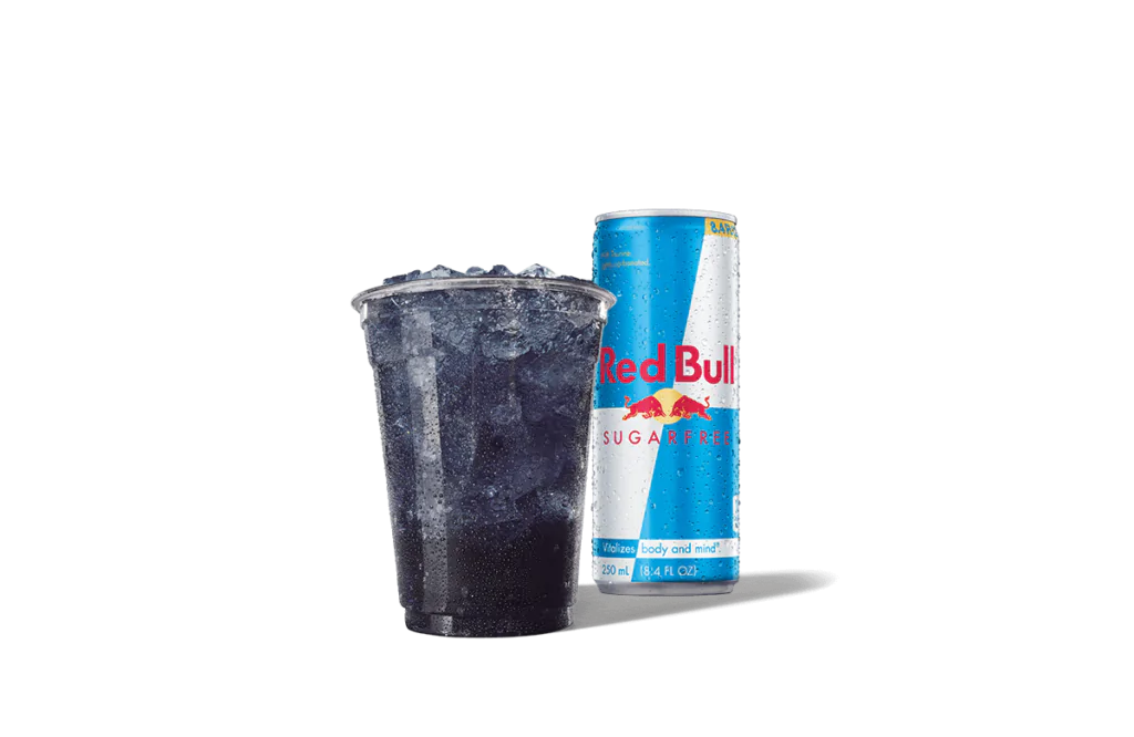 A Berry Purple Daze Red Bull® Sugarfree Infusion drink served in a clear cup filled with ice, highlighting its vibrant purple hue. Accompanying the drink is a can of Red Bull® Sugarfree, featuring the iconic blue and silver design with "Sugarfree" written in bold red letters. The can is covered in droplets, emphasizing its chilled and refreshing nature.
