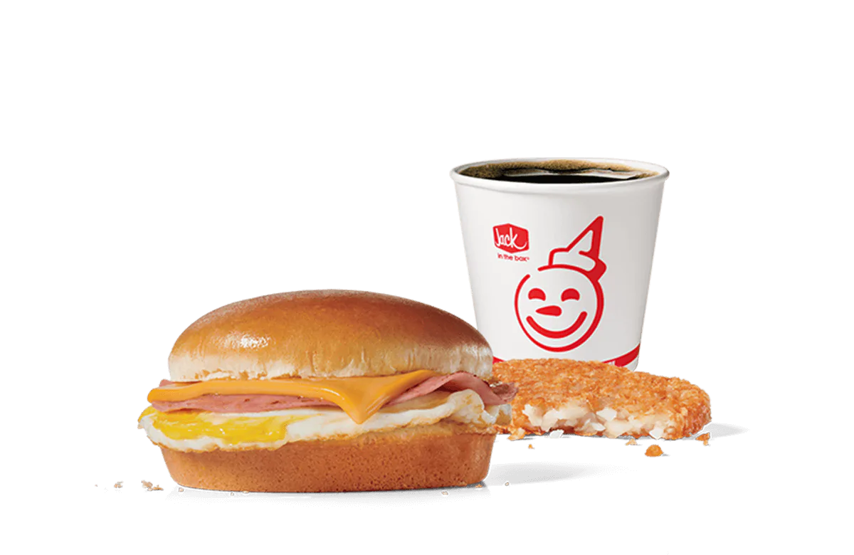 A Breakfast Jack® Combo from Jack in the Box featuring a classic sandwich with ham, egg, and American cheese on a toasted bun, alongside a crispy hash brown and a cup of hot coffee. The set is displayed against a transparent background.