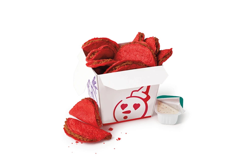 A box of classic spicy tiny tacos from Jack in the Box, featuring mini tacos with a bright red, spicy coating. The tacos are served in a white box with red and purple designs, including a heart-eyed character illustration. A few tacos are displayed outside the box alongside a cup of creamy dipping sauce.