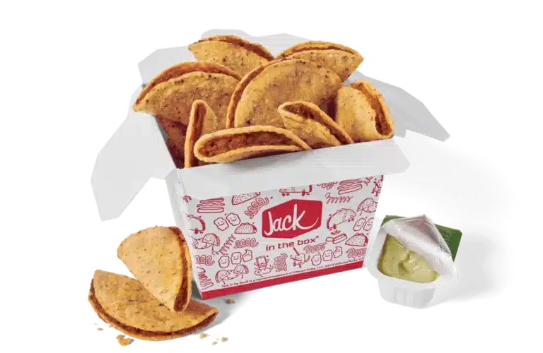 A box of classic tiny tacos from Jack in the Box, featuring small, crispy taco shells filled with seasoned beef. The tacos are presented in a branded red and white box with a design of playful illustrations. A container of green avocado lime sauce, partially opened, is placed beside the box, and a few tacos are scattered in front.