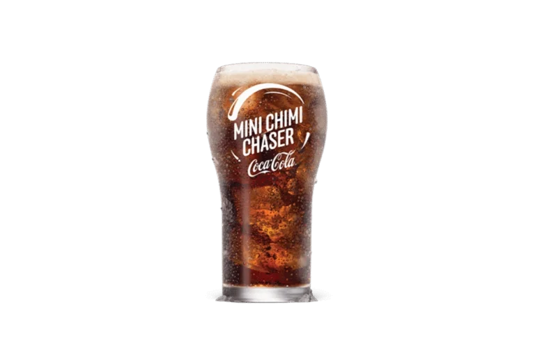 A glass of Coca-Cola® Mini Chimi Chaser with visible carbonation and foam on top. The glass features the Coca-Cola® logo and "Mini Chimi Chaser" text in white, emphasizing its refreshing and fizzy appearance. The dark background enhances the drink's vibrant, bubbly texture.