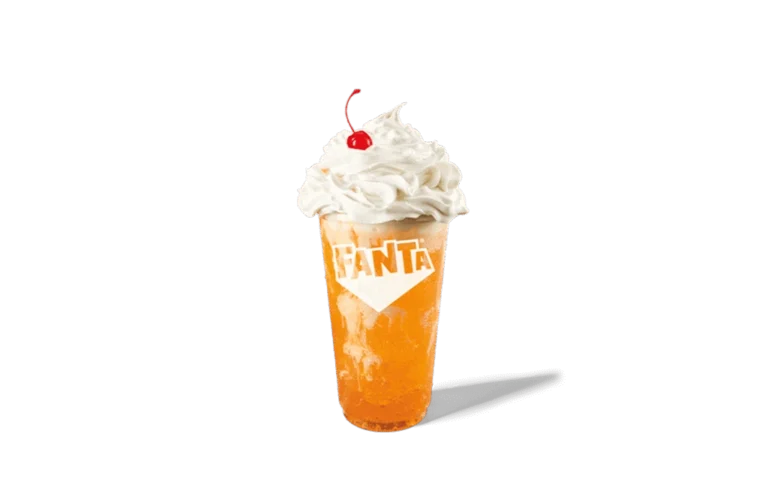A glass of Fanta® Orange Swirl Twisted Soda with a bright orange soda base, swirled with creamy textures. Topped with a generous dollop of whipped cream and a cherry, the drink offers a playful and refreshing appeal. The Fanta® logo is prominently displayed on the glass, adding brand visibility against transparent background.