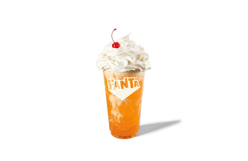 A glass of Fanta® Orange Swirl Twisted Soda with a bright orange soda base, swirled with creamy textures. Topped with a generous dollop of whipped cream and a cherry, the drink offers a playful and refreshing appeal. The Fanta® logo is prominently displayed on the glass, adding brand visibility against transparent background.