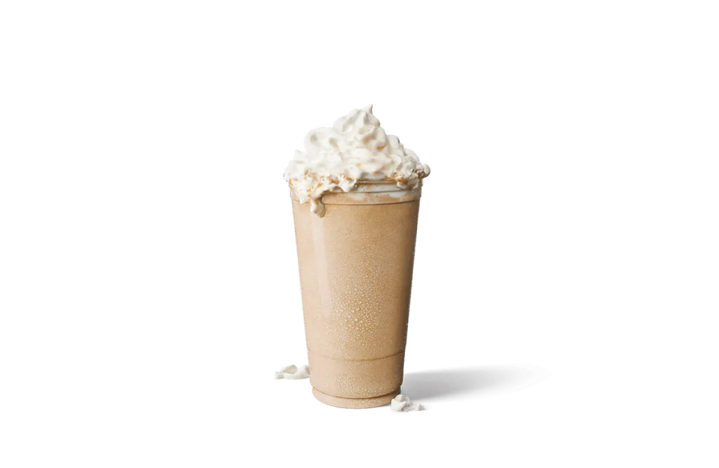 A tall glass of iced vanilla creamaccino, showcasing a creamy light beige color and topped with a generous swirl of whipped cream. The glass is frosty with condensation, highlighting the drink's chilled and refreshing nature, with a hint of spilled whipped cream nearby for an artistic touch.