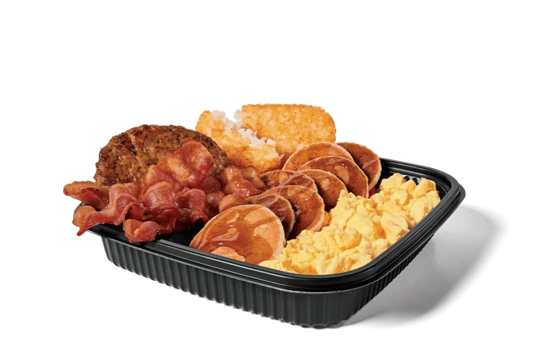 Image of a jumbo breakfast platter featuring crispy bacon strips, two sausage patties, fluffy scrambled eggs, golden hash browns, and mini pancakes drizzled with syrup, all served in a black plastic tray.