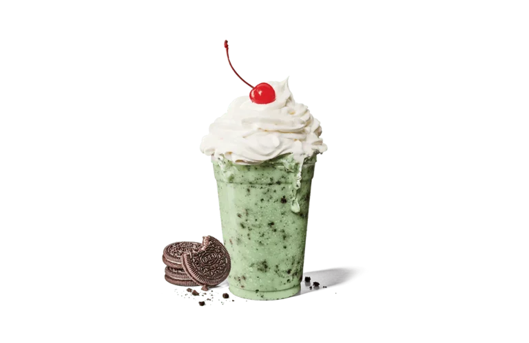 A refreshing Oreo® cookie mint shake in a clear cup, topped with a generous amount of whipped cream and a single cherry. The shake has a vibrant mint green color and is speckled with crushed Oreo® cookie pieces. Several whole Oreo® cookies lie beside the cup transparent background, some crumbled, adding to the dessert's indulgent appeal.