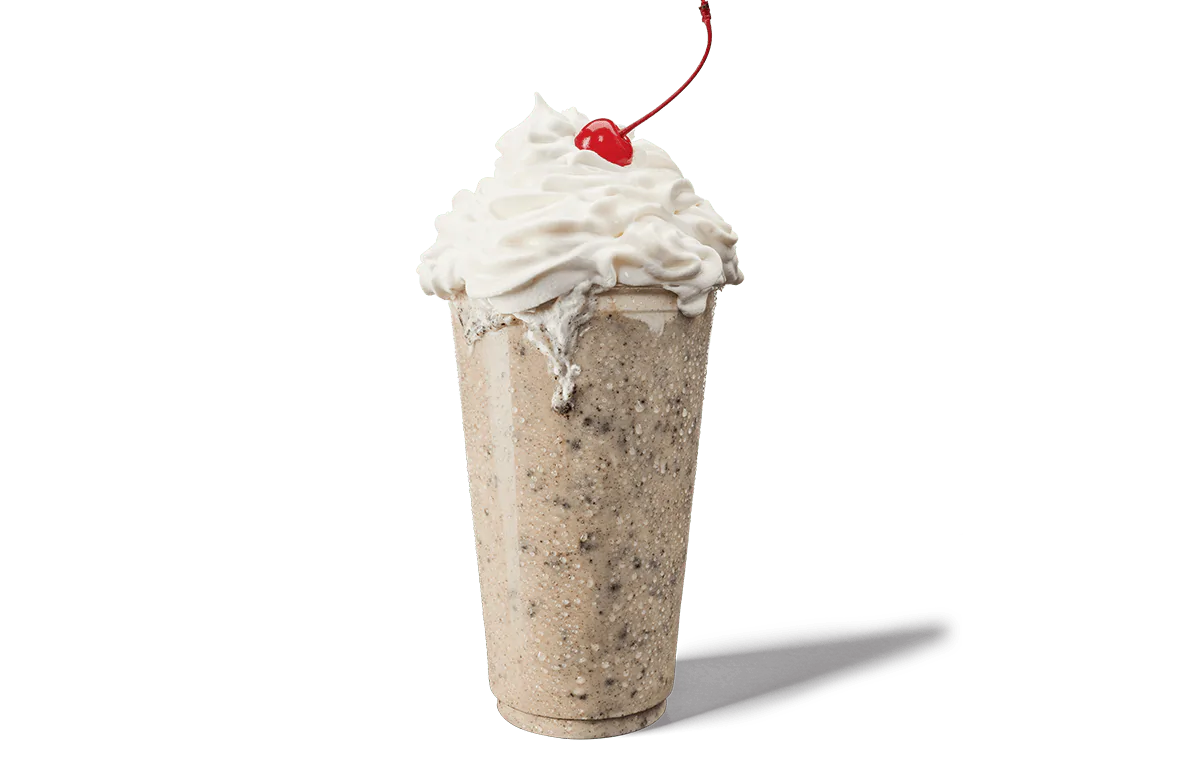A tall, creamy Oreo® shake overflowing with whipped cream, topped with a single bright red cherry. The shake is speckled with pieces of Oreo® cookies throughout, giving it a textured look against transparent background. The whipped cream is generously swirled on top, creating an appealing and indulgent dessert.