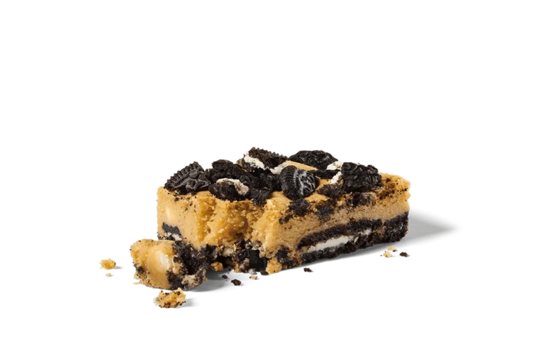 A decadent slice of Oreo® Ultimate Cookie Bar on transparent background, featuring layers of creamy filling and Oreo® cookie pieces. The top is generously sprinkled with crumbled Oreo® cookies, adding a rich texture and deep chocolate flavor to the dessert. The bar is showcased with crumbs scattered around, highlighting its freshly cut and indulgent appearance.