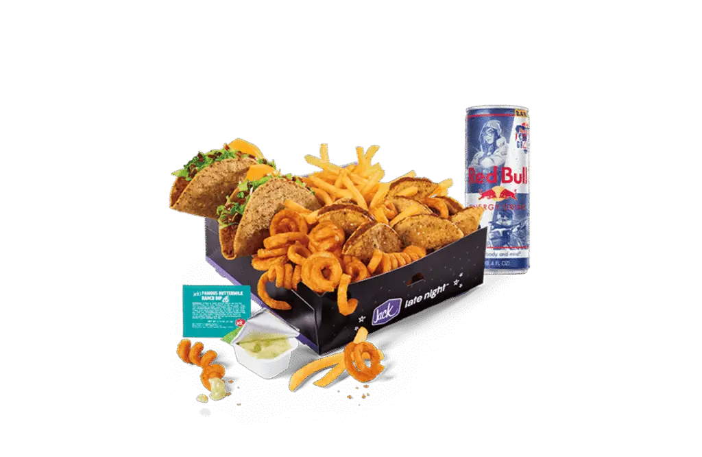 A Red Bull Gaming Box from Jack in the Box, featuring an assortment of snacks including crunchy tacos, golden fries, tiny tacos, and curly fries. The items are neatly arranged in a black box labeled "Jack late night" with stars and a moon design. A can of Red Bull Energy Drink is placed next to the box, along with a dipping sauce container and packet on the side. A few curly fries and tiny tacos are scattered around for a casual presentation.