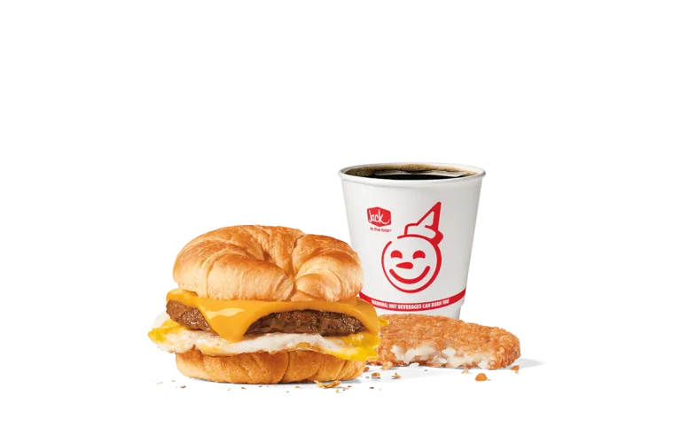 Image of the Sausage Croissant Combo from Jack in the Box, featuring a breakfast sandwich with a savory sausage patty, fried egg, and melted cheese nestled inside a flaky croissant. Accompanied by a hash brown and a cup of coffee, all set against a transparent background with the iconic smiling Jack logo.