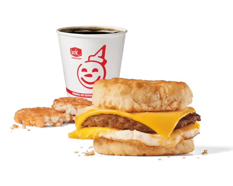 A hearty breakfast combo featuring a biscuit sandwich with a savory sausage patty, melted cheese, and a fried egg, served with a hash brown patty and a cup of hot coffee in a Jack in the Box branded cup. The background is transparent, emphasizing the meal.