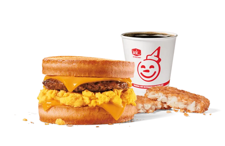 Image of Jack in the Box's Sausage, Egg & Cheese Scrambler Combo, featuring a breakfast sandwich with a sausage patty, scrambled eggs, and melted cheese on a muffin, alongside a hash brown and a coffee cup with the iconic smiling face logo. The backdrop is transparent, focusing attention on the rich colors and textures of the meal components.