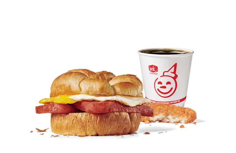A promotional image featuring Jack in the Box's breakfast combo with a croissant sandwich containing Spam, egg, and cheese, paired with a hash brown and a coffee in a cup adorned with the Jack in the Box logo. The meal is presented on a transparent background, emphasizing the food items.