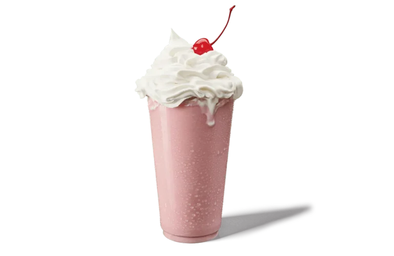 A strawberry shake in a tall glass, topped with an abundant swirl of whipped cream and a bright red cherry. The shake is a soft pink color, with visible condensation on the glass, suggesting a cold and refreshing beverage. The whipped cream overflows slightly, adding to the indulgent look of this classic dessert.