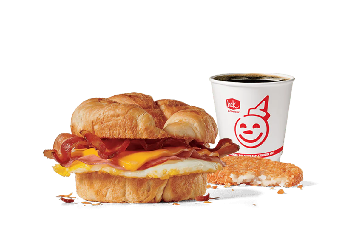 Image of the Supreme Croissant Combo from Jack in the Box, featuring a breakfast sandwich with crispy bacon, a fried egg, and melty cheese inside a flaky croissant. Accompanied by a hash brown and a cup of coffee, all set against transparent background with the iconic Jack in the Box logo.