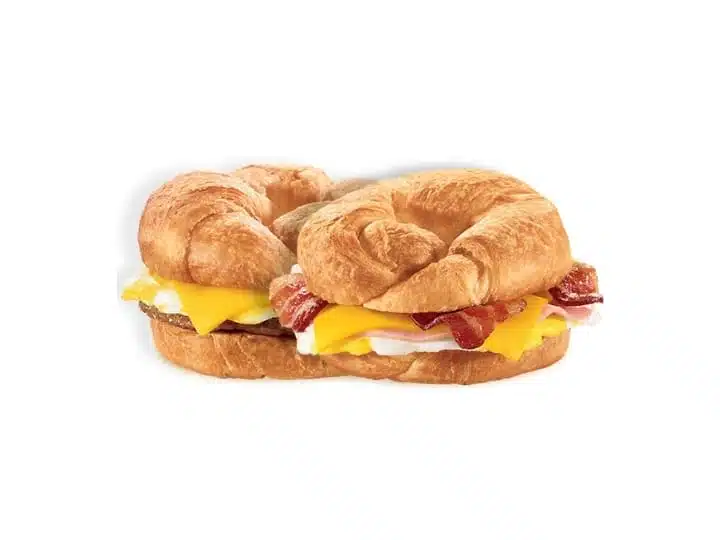 A freshly prepared breakfast croissant sandwich featuring flaky, golden-brown pastry layers with crispy bacon, a slice of melted yellow cheese, and a fried egg visible within, all set on a white background.
