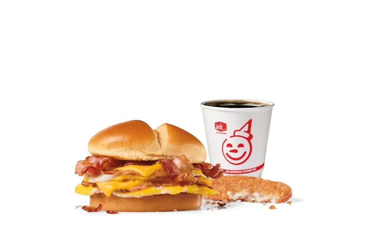 A delectable Ultimate Breakfast Sandwich Combo from Jack in the Box featuring a sandwich with layers of bacon, ham, egg, and melted American cheese on a toasted bun. This combo includes a crispy hash brown and a hot coffee in a branded cup, all against a transparent background.