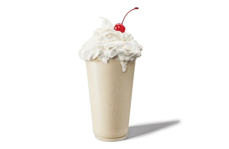 A classic vanilla milkshake in a tall glass, overflowing with whipped cream and topped with a single bright red cherry. The glass shows a refreshing condensation and the background is transparent, enhancing the simplicity and appeal of the milkshake.