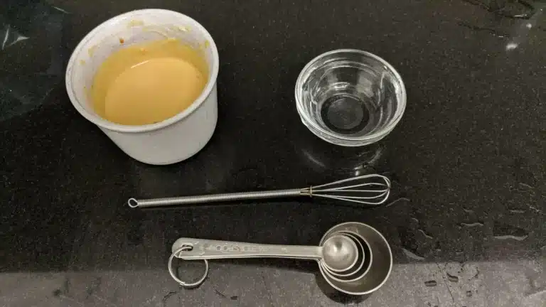 A small white container filled with a creamy, orange-colored sauce sits on a black countertop. Next to it are a small glass bowl, a mini whisk, and a set of stainless steel measuring spoons. The countertop has some water droplets, suggesting recent use in food preparation.